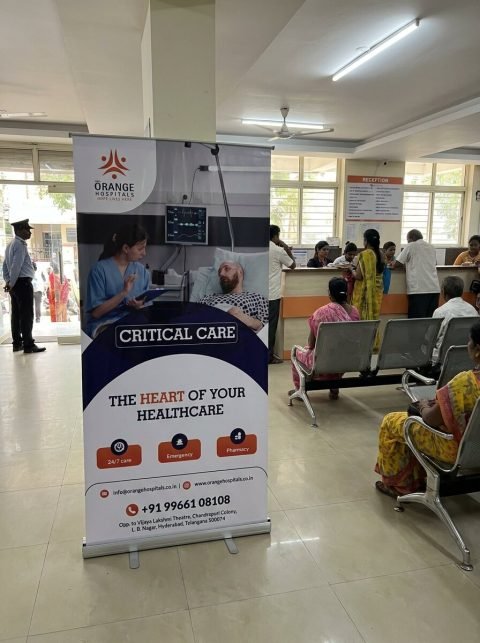 Orange Hospitals critical care roll-up standee in a busy hospital lobby near reception, photo mockup
